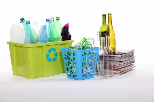 A lime green plastic recycling bin with a blue recycling symbol on the side is positioned in the foreground, filled with various plastic bottles and containers in shades of clear, green, and blue. To its right, a smaller blue plastic basket with circular cutouts holds additional plastic bottles. Behind these containers, there are several empty glass bottles in green, yellow, and clear glass, with smooth surfaces and some with narrow necks, standing upright on a clean, white surface. Adjacent to the glass bottles, a neatly stacked pile of newspapers or paper documents is present, with visible pages and slightly crumpled edges. The background is plain white, emphasizing the arrangement of waste materials suitable for a rubbish removal or recycling service. The scene is well-lit with even lighting, highlighting the textures of the plastic and glass objects, aligning with the context of rubbish collection activities, particularly in the Thornton Heath area.