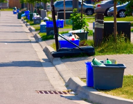 A grey plastic storage bin placed on the grassy verge alongside a sidewalk, containing various discarded plastic items such as bottles and containers, with a blue recycling bin nearby filled with similar waste. Close to the curb, there is a dark blue push broom with a long handle leaning against the curb. Further along the sidewalk, multiple blue recycling bins and black waste bins are lined up on the pavement, each filled with rubbish, and there are additional waste containers scattered along the residential street. The street is bordered by small trees and neatly kept grass, with parked cars visible in the background. The overall scene suggests a typical street in Thornton Heath, with waste collection and rubbish management in progress, illustrating the context of local rubbish disposal services provided by Garden Clearance Thornton Heath.