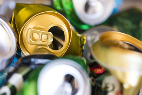 A close-up view of a pile of crushed aluminium and steel cans, including a prominent yellow-green can in the foreground with an open tab, surrounded by other cans in various shades of colors such as silver, green, white, and gold. The cans exhibit metallic textures with reflective surfaces and slight dents from compression. They are stacked closely together, some overlapping, on what appears to be an outdoor surface, possibly a driveway or street in Thornton Heath, London. The background is blurred, drawing attention to the details of the cans and emphasizing their discarded state. This image visually relates to rubbish and waste collection services offered by Garden Clearance Thornton Heath, highlighting the importance of proper disposal of household metals and cans as part of rubbish removal or garden clearance projects.