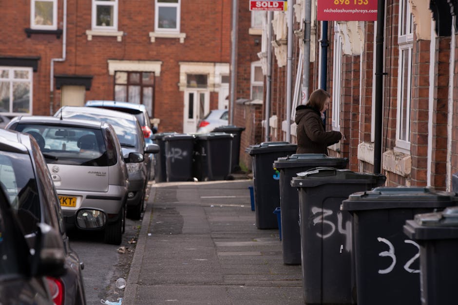 A black wheelie bin with the label 'ST. JOHN'S' is positioned on the pavement beside the curb on a quiet street at night. The bin is overflowing with mixed waste materials, including cardboard boxes,
