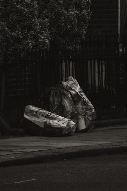 Three large plastic sacks filled with waste material are placed on the pavement near a wooden fence and a tree with dense foliage. The sacks appear to contain household rubbish or discarded items, wit